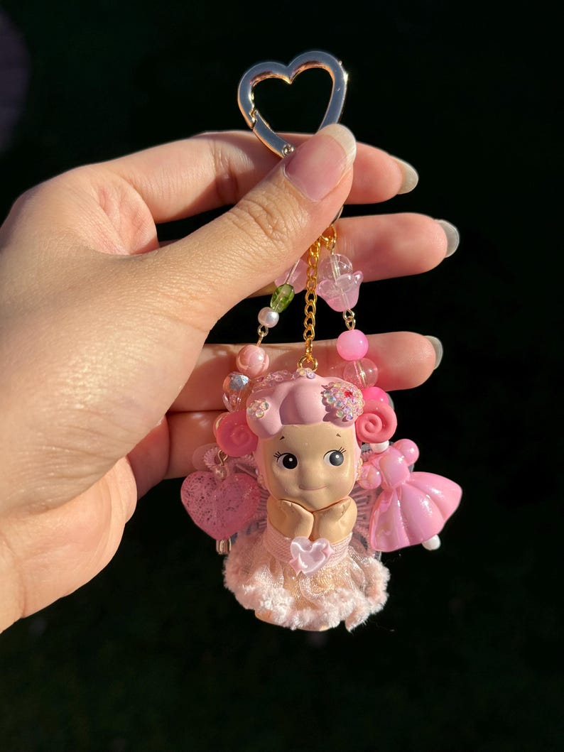 May include: A pink and gold keychain featuring a small doll with a heart-shaped charm. The doll is wearing a pink dress and has pink hair. The keychain also includes pink beads and a gold heart-shaped clasp.