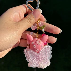 May include: Pink keychain featuring a pig-like character with a butterfly-shaped design. The keychain has a gold heart-shaped clasp and is adorned with pink and clear beads, pearls, and a gold chain. The character has flower details.