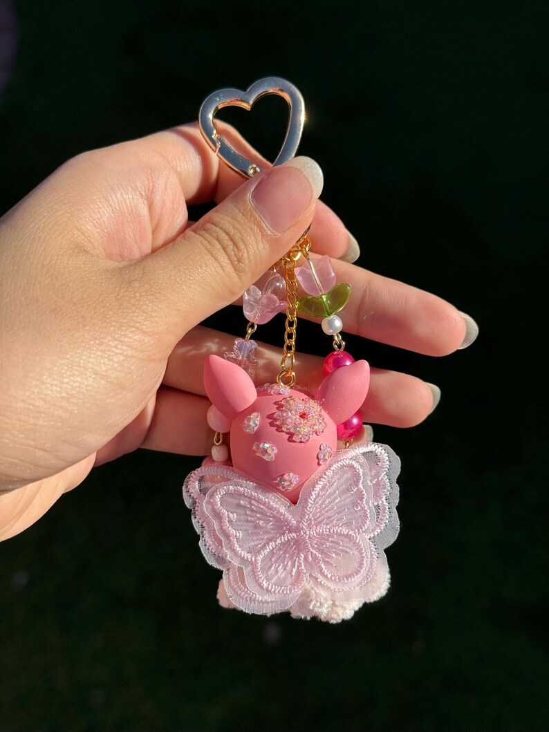 May include: Pink keychain featuring a pig-like character with a butterfly-shaped design. The keychain has a gold heart-shaped clasp and is adorned with pink and clear beads, pearls, and a gold chain. The character has flower details.