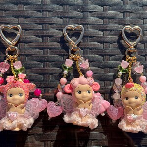 May include: Three keychains featuring fairy figurines with pink wings and dresses. Each keychain has a gold heart-shaped clasp and is adorned with pink beads, flowers, and heart charms. The fairies have rosy cheeks and are in a seated position.