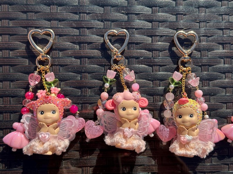 May include: Three keychains featuring fairy figurines with pink wings and dresses. Each keychain has a gold heart-shaped clasp and is adorned with pink beads, flowers, and heart charms. The fairies have rosy cheeks and are in a seated position.