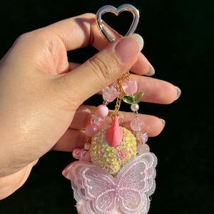 May include: A decorative keychain with a gold heart-shaped clasp. The keychain features pink beads, a butterfly, and a cluster of pink and yellow decorative elements. The butterfly has a delicate, lacy design.
