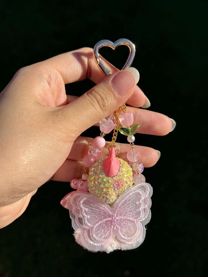 May include: A decorative keychain with a gold heart-shaped clasp. The keychain features pink beads, a butterfly, and a cluster of pink and yellow decorative elements. The butterfly has a delicate, lacy design.