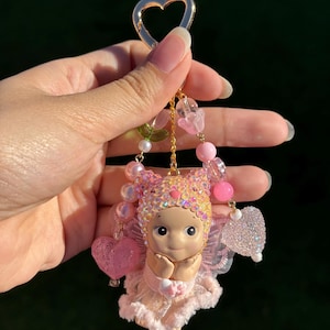 May include: A pink and gold keychain featuring a small doll with a heart-shaped charm. The doll has a pink dress, wings, and a beaded headpiece. The keychain includes pink and clear beads and a gold heart-shaped clasp.