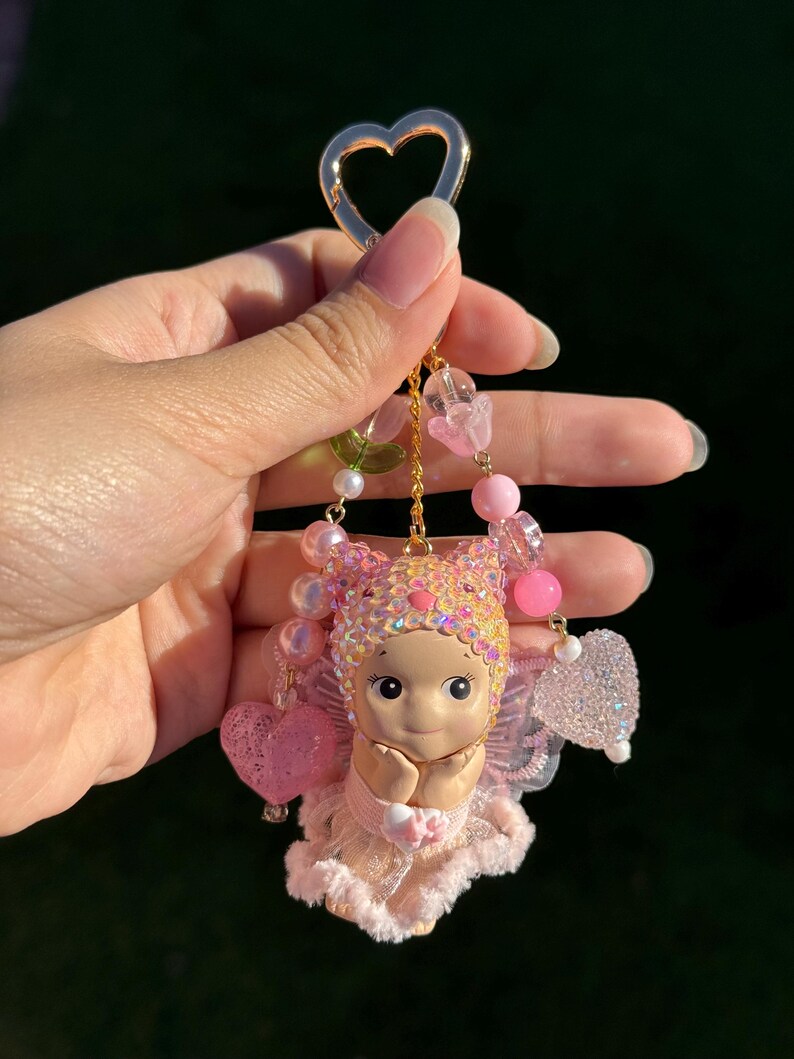 May include: A pink and gold keychain featuring a small doll with a heart-shaped charm. The doll has a pink dress, wings, and a beaded headpiece. The keychain includes pink and clear beads and a gold heart-shaped clasp.