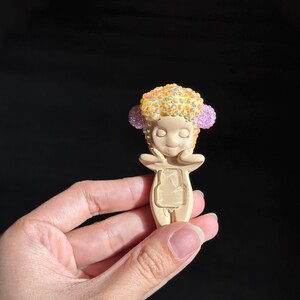 May include: A small, beige figurine featuring a stylized character with closed eyes and hands touching its cheeks. The character has a textured, rounded head covered in iridescent yellow and pink rhinestones, and a rectangular cutout on its torso.
