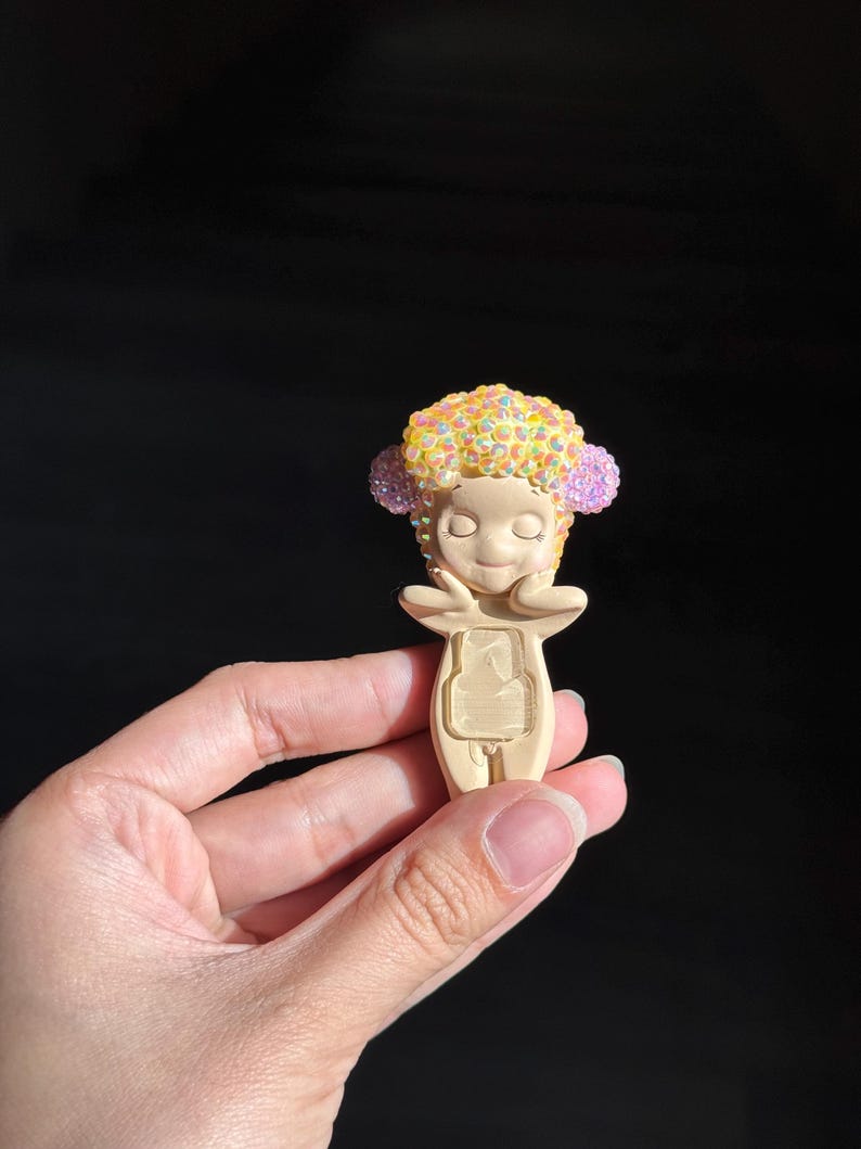 May include: A small, beige figurine featuring a stylized character with closed eyes and hands touching its cheeks. The character has a textured, rounded head covered in iridescent yellow and pink rhinestones, and a rectangular cutout on its torso.