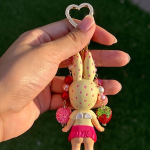 May include: A keychain featuring a pale yellow rabbit figure with pink spots, small white wings, and a bright pink skirt. The figure is adorned with dangling charms including red and white beads, a pink glittery sphere, and a green and red strawberry.
