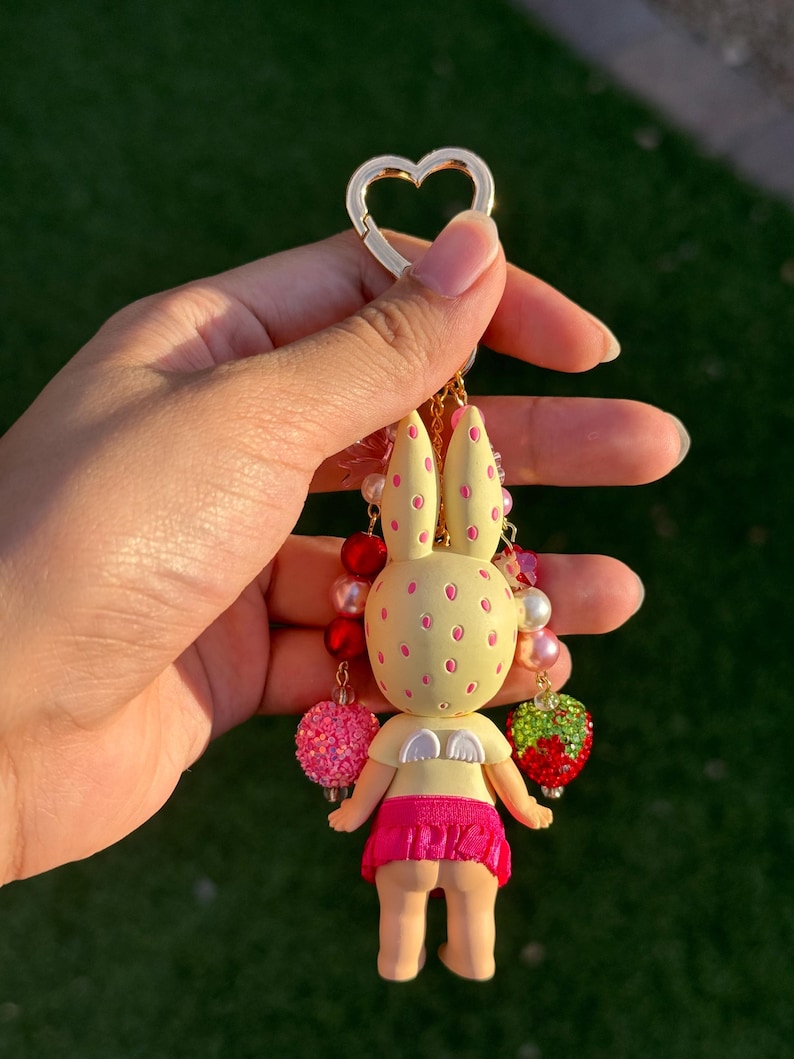 May include: A keychain featuring a pale yellow rabbit figure with pink spots, small white wings, and a bright pink skirt. The figure is adorned with dangling charms including red and white beads, a pink glittery sphere, and a green and red strawberry.