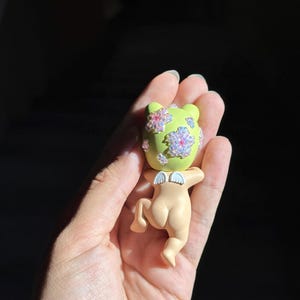 May include: A small figurine featuring a light green head adorned with iridescent, flower-shaped embellishments and a peach-colored body with small white wings. The character is depicted in a crawling position, showcasing a smooth, matte finish.
