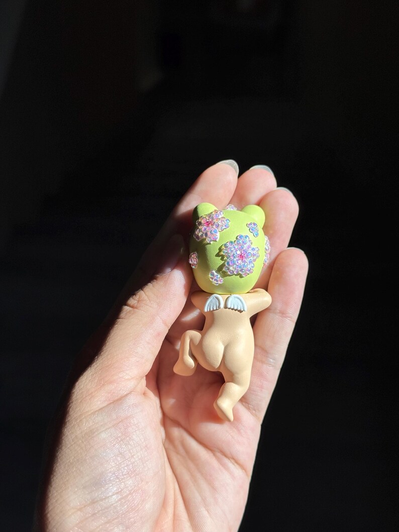 May include: A small figurine featuring a light green head adorned with iridescent, flower-shaped embellishments and a peach-colored body with small white wings. The character is depicted in a crawling position, showcasing a smooth, matte finish.