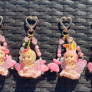 May include: Six keychains with heart-shaped clasps, featuring small angel figurines in pink dresses and wings. Each keychain is adorned with pink beads and floral accents. One figurine has bunny ears.
