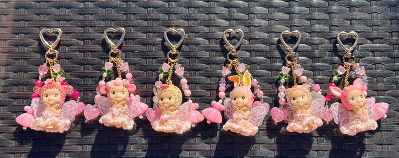May include: Six keychains with heart-shaped clasps, featuring small angel figurines in pink dresses and wings. Each keychain is adorned with pink beads and floral accents. One figurine has bunny ears.