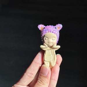 May include: A small, beige figurine featuring a character with closed eyes and hands touching its cheeks. The character wears a head covering adorned with numerous small, iridescent, light purple and pink crystals that catch the light.