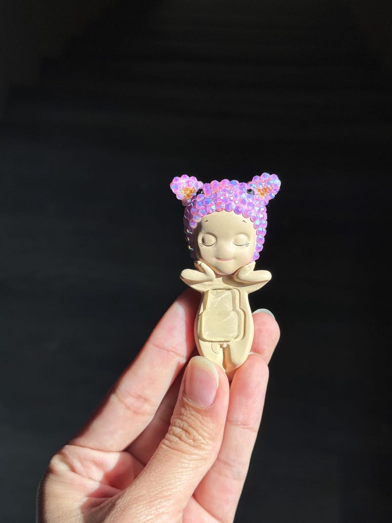 May include: A small, beige figurine featuring a character with closed eyes and hands touching its cheeks. The character wears a head covering adorned with numerous small, iridescent, light purple and pink crystals that catch the light.