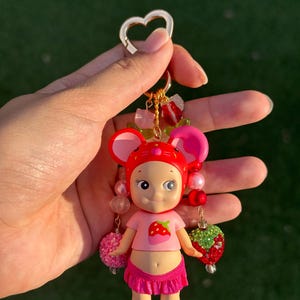 May include: A keychain featuring a small doll figure wearing a red strawberry-themed hood and a pink shirt with a strawberry graphic. The doll has a pink ruffled skirt and is decorated with dangling beads, including pink spheres and a red strawberry charm.