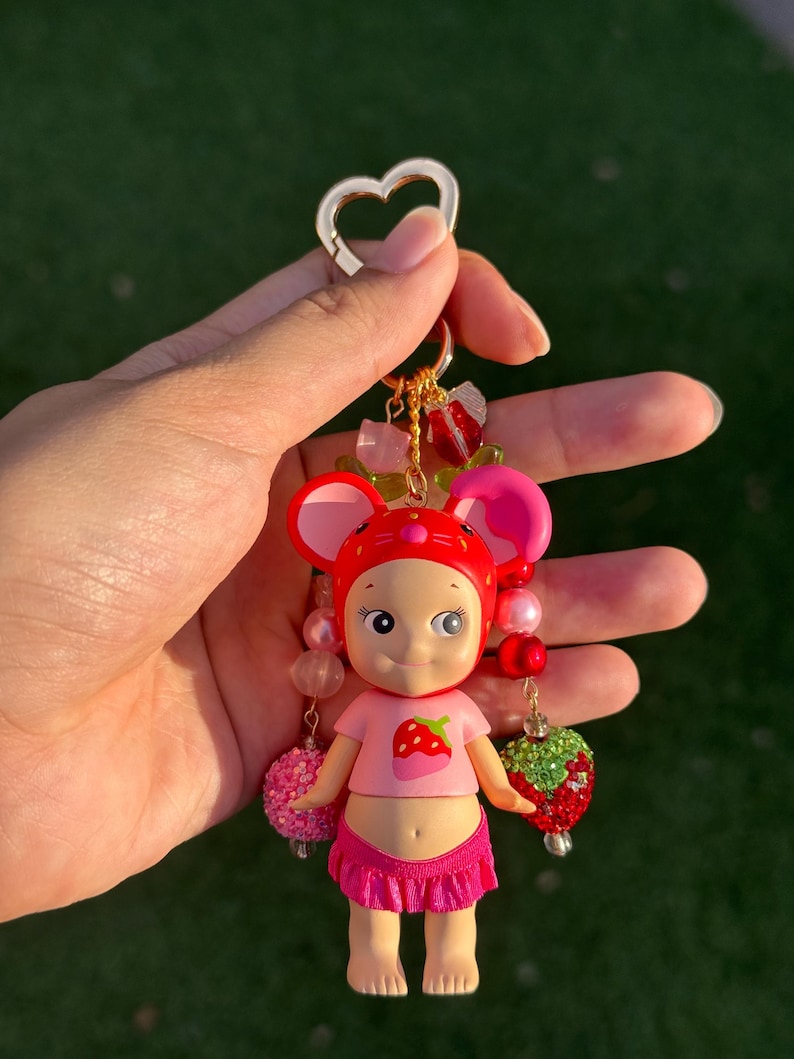 May include: A keychain featuring a small doll figure wearing a red strawberry-themed hood and a pink shirt with a strawberry graphic. The doll has a pink ruffled skirt and is decorated with dangling beads, including pink spheres and a red strawberry charm.