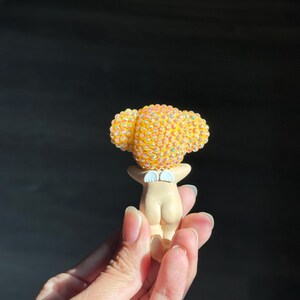 May include: A small figurine featuring a smooth, light-toned body with a pair of white wings on the back. The head is covered in a dense cluster of iridescent, yellow-orange spherical beads that catch the light, creating a textured appearance.