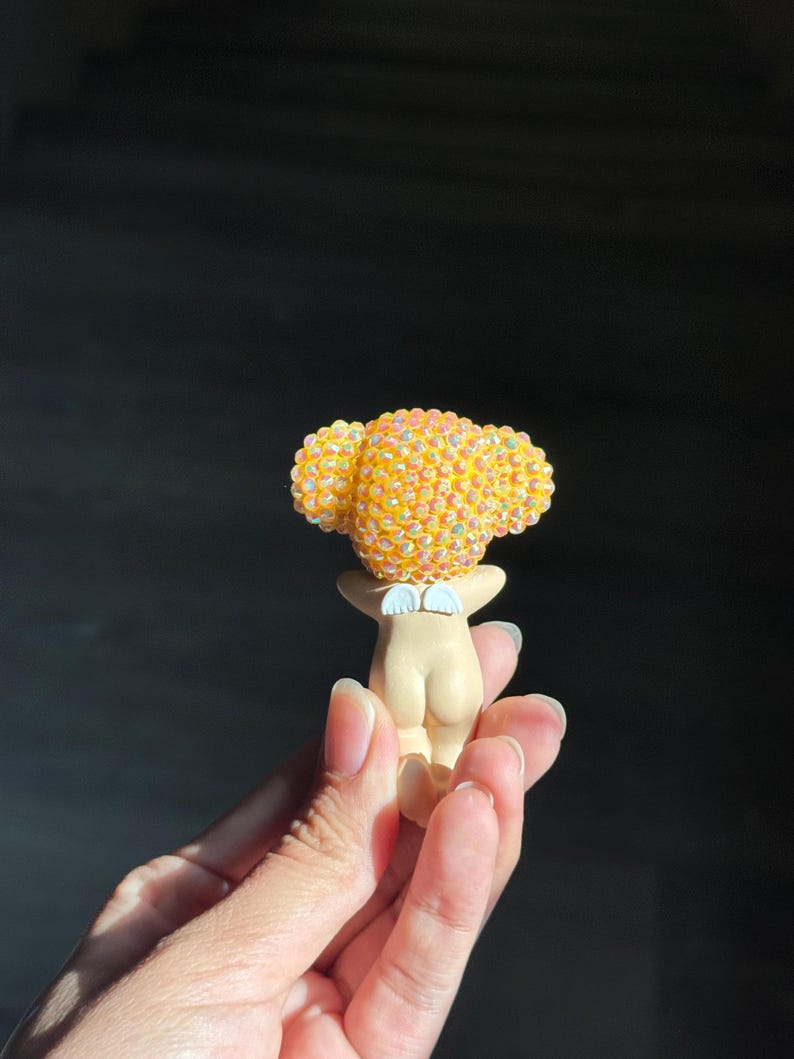 May include: A small figurine featuring a smooth, light-toned body with a pair of white wings on the back. The head is covered in a dense cluster of iridescent, yellow-orange spherical beads that catch the light, creating a textured appearance.