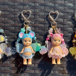 Puede incluir: Cuatro llaveros con cierres en forma de coraz&oacute;n, cada uno con una peque&ntilde;a mu&ntilde;eca con alas y un sombrero con tem&aacute;tica de animales. Las mu&ntilde;ecas son de varios colores, incluyendo gris, azul, rosa y amarillo, con detalles de cuentas decorativas.