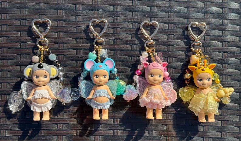 Puede incluir: Cuatro llaveros con cierres en forma de coraz&oacute;n, cada uno con una peque&ntilde;a mu&ntilde;eca con alas y un sombrero con tem&aacute;tica de animales. Las mu&ntilde;ecas son de varios colores, incluyendo gris, azul, rosa y amarillo, con detalles de cuentas decorativas.
