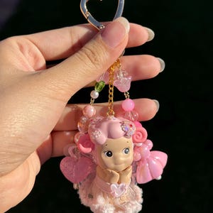 May include: A pink keychain featuring a figurine of a smiling character with pink hair and a dress. The keychain has a gold heart-shaped clasp and is adorned with pink beads and charms. The character is holding a heart.