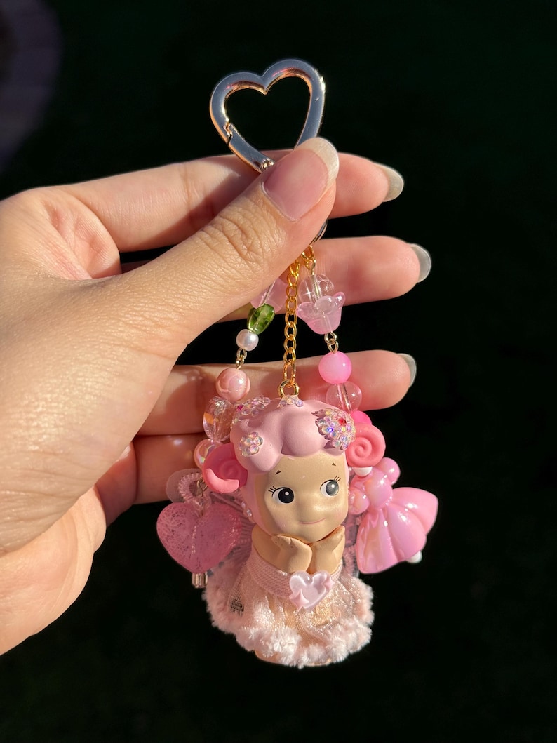 May include: A pink keychain featuring a figurine of a smiling character with pink hair and a dress. The keychain has a gold heart-shaped clasp and is adorned with pink beads and charms. The character is holding a heart.