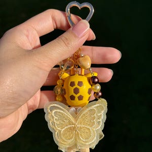 Puede incluir: Un llavero amarillo y marr&oacute;n con forma de jirafa con un cierre dorado en forma de coraz&oacute;n. El llavero presenta un dise&ntilde;o de mariposa hecho de encaje amarillo y est&aacute; adornado con cuentas amarillas y marrones.