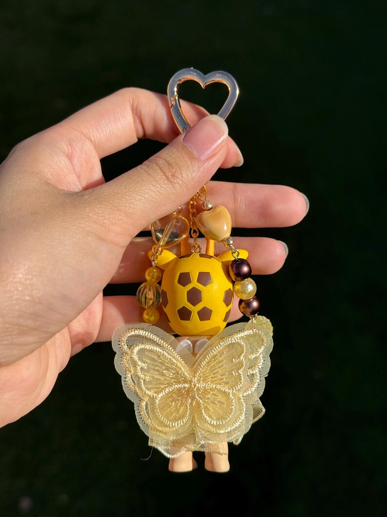 Puede incluir: Un llavero amarillo y marr&oacute;n con forma de jirafa con un cierre dorado en forma de coraz&oacute;n. El llavero presenta un dise&ntilde;o de mariposa hecho de encaje amarillo y est&aacute; adornado con cuentas amarillas y marrones.