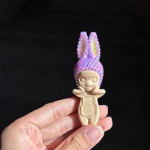 May include: A small figurine with a smooth beige body and a head covered in iridescent purple crystals shaped like rabbit ears. The figure has closed eyes, a small nose, and hands resting near its cheeks, with a rectangular indentation on its torso.