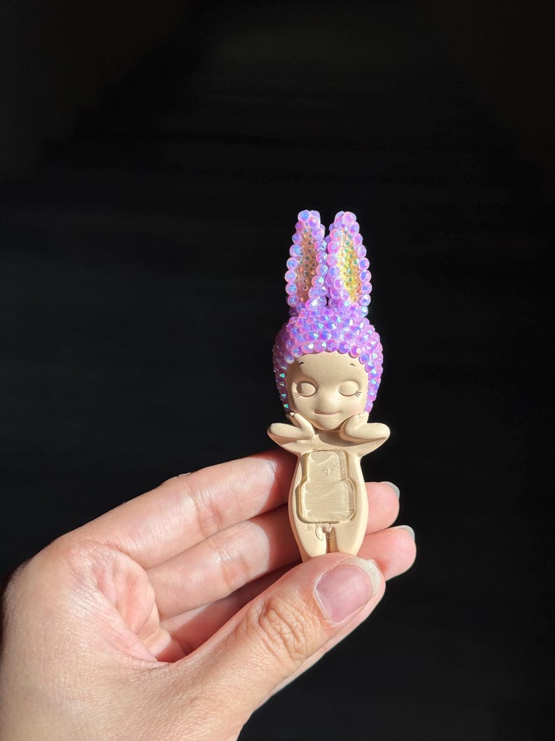 May include: A small figurine with a smooth beige body and a head covered in iridescent purple crystals shaped like rabbit ears. The figure has closed eyes, a small nose, and hands resting near its cheeks, with a rectangular indentation on its torso.