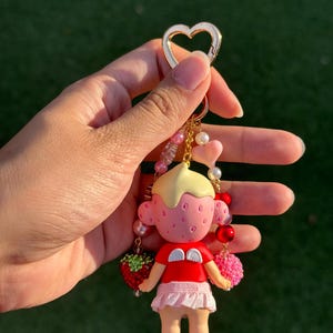 May include: A keychain featuring a small figurine with a strawberry-textured head, blonde hair, and a red shirt with white wings. The figure wears a light pink skirt and is attached to a gold heart-shaped clasp with dangling beads in pink, red, and white.