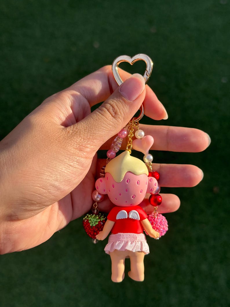 May include: A keychain featuring a small figurine with a strawberry-textured head, blonde hair, and a red shirt with white wings. The figure wears a light pink skirt and is attached to a gold heart-shaped clasp with dangling beads in pink, red, and white.