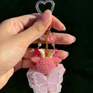 May include: A pink and gold keychain with a heart-shaped clasp. The keychain features a beaded goat figure with small horns, a butterfly, and dangling heart charms. The butterfly is made of pink lace.
