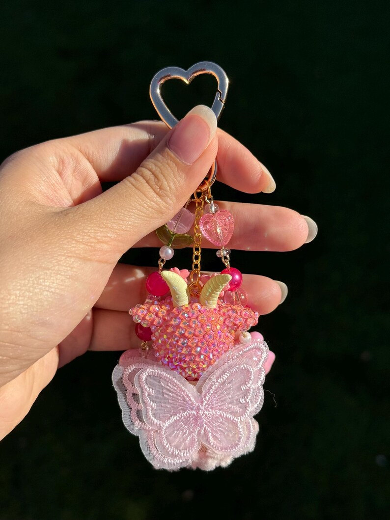 May include: A pink and gold keychain with a heart-shaped clasp. The keychain features a beaded goat figure with small horns, a butterfly, and dangling heart charms. The butterfly is made of pink lace.