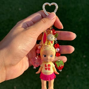 May include: A keychain featuring a small figurine with yellow rabbit ears, wearing a cream-colored top with the red text LOVE and a bright pink ruffled skirt. The charm is accented with red and pink beads, a heart-shaped bead, and a strawberry charm.