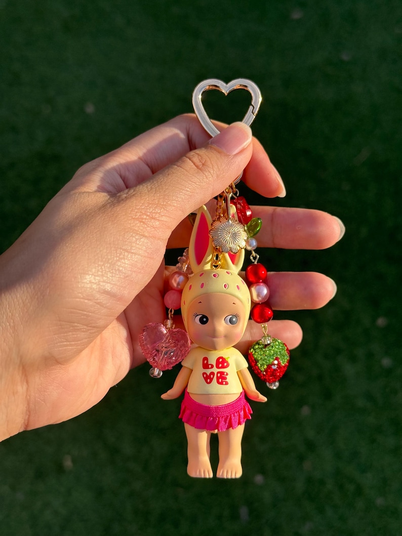 May include: A keychain featuring a small figurine with yellow rabbit ears, wearing a cream-colored top with the red text LOVE and a bright pink ruffled skirt. The charm is accented with red and pink beads, a heart-shaped bead, and a strawberry charm.