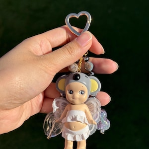 Puede incluir: Un peque&ntilde;o llavero con forma de figura con un gorro de koala y alas de hada. La figura lleva un top y una parte inferior con volantes blancos. El llavero tiene un cierre dorado en forma de coraz&oacute;n y una cadena de cuentas.