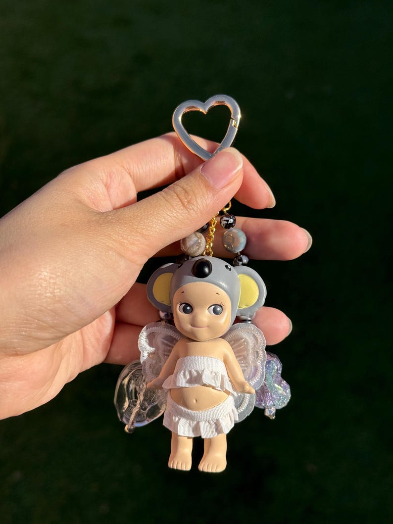 Puede incluir: Un peque&ntilde;o llavero con forma de figura con un gorro de koala y alas de hada. La figura lleva un top y una parte inferior con volantes blancos. El llavero tiene un cierre dorado en forma de coraz&oacute;n y una cadena de cuentas.