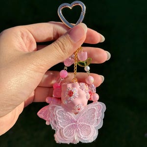 May include: A pink keychain with a gold heart-shaped clasp. The keychain features a pink figure with a butterfly design, adorned with pink beads and floral accents. The figure is suspended from a gold chain.