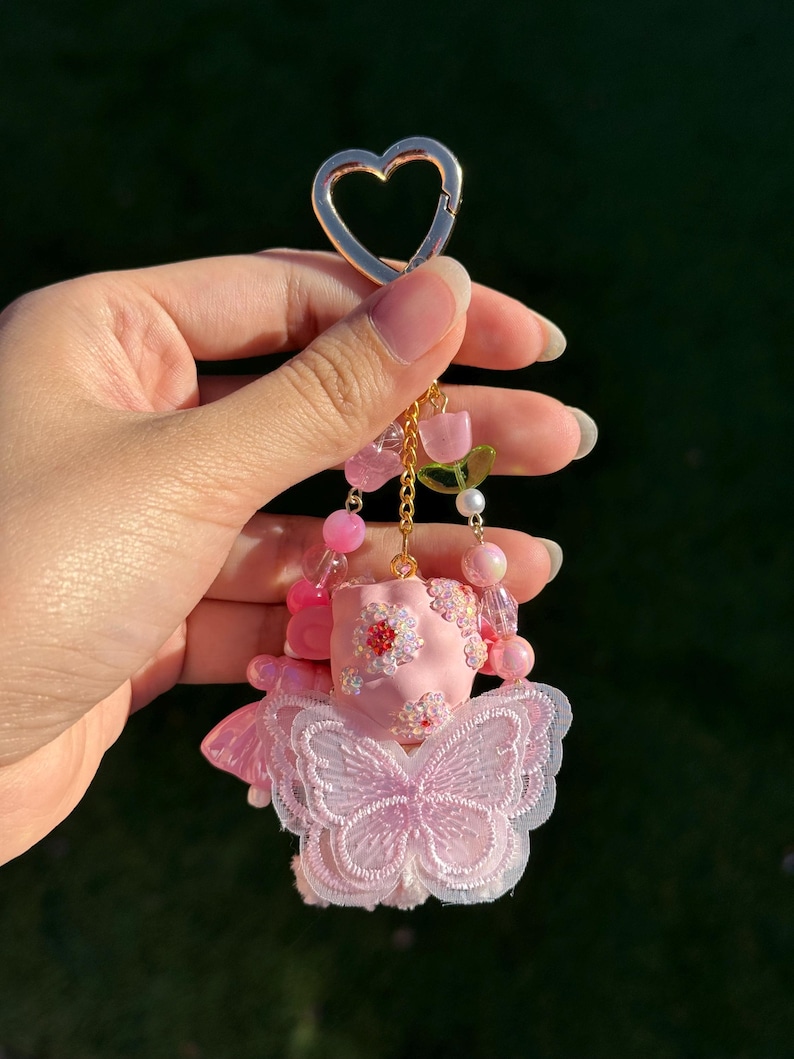 May include: A pink keychain with a gold heart-shaped clasp. The keychain features a pink figure with a butterfly design, adorned with pink beads and floral accents. The figure is suspended from a gold chain.