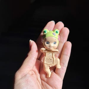 May include: A small figurine of a baby wearing a light green frog hat with pink floral accents. The figure has a light skin tone, large dark eyes, and a small smile. Embossed text on the chest reads Sonny Angel, Dreams, and China.
