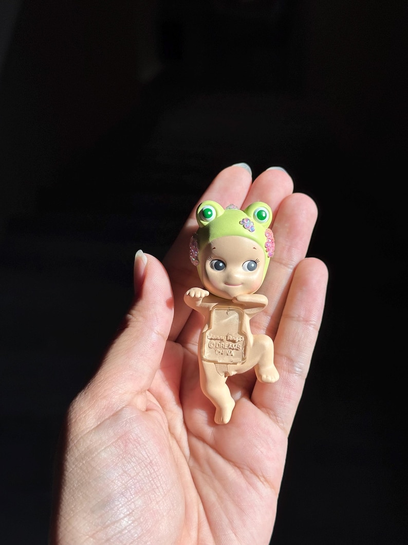 May include: A small figurine of a baby wearing a light green frog hat with pink floral accents. The figure has a light skin tone, large dark eyes, and a small smile. Embossed text on the chest reads Sonny Angel, Dreams, and China.
