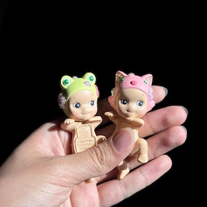 May include: Two small figurines with light skin tones, one wearing a lime green frog hood and the other a pink cat hood. Both figures feature small, iridescent, multi-colored crystal embellishments on the sides of their hoods and faces.