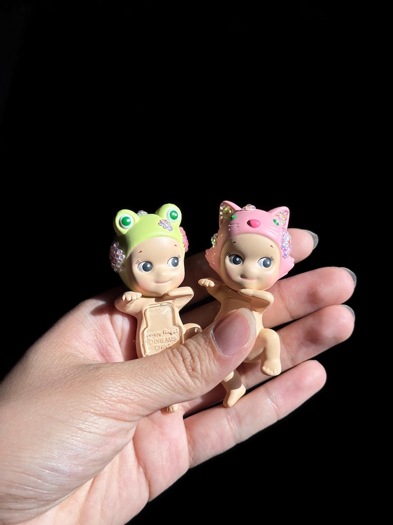 May include: Two small figurines with light skin tones, one wearing a lime green frog hood and the other a pink cat hood. Both figures feature small, iridescent, multi-colored crystal embellishments on the sides of their hoods and faces.