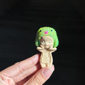 May include: A small, beige figurine featuring a stylized character with closed eyes and hands touching its cheeks. The character wears a bright lime green, textured cap adorned with iridescent sequins and a single small pink flower at the center.