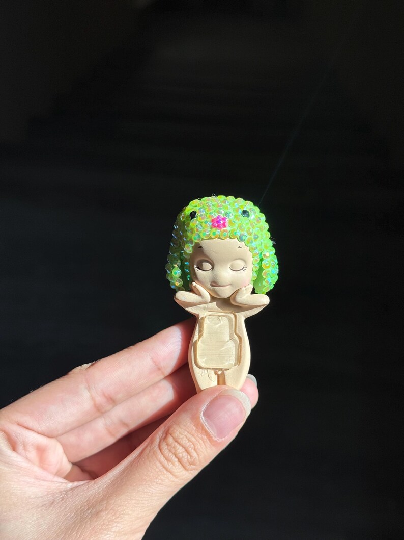 May include: A small, beige figurine featuring a stylized character with closed eyes and hands touching its cheeks. The character wears a bright lime green, textured cap adorned with iridescent sequins and a single small pink flower at the center.