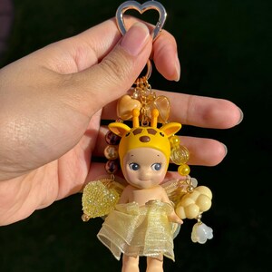 Puede incluir: Un llavero amarillo y dorado con una peque&ntilde;a mu&ntilde;eca vestida de jirafa. La mu&ntilde;eca tiene una capucha de jirafa amarilla, una falda amarilla transparente y peque&ntilde;as alas amarillas. El llavero incluye cuentas doradas y un cierre en forma de coraz&oacute;n.