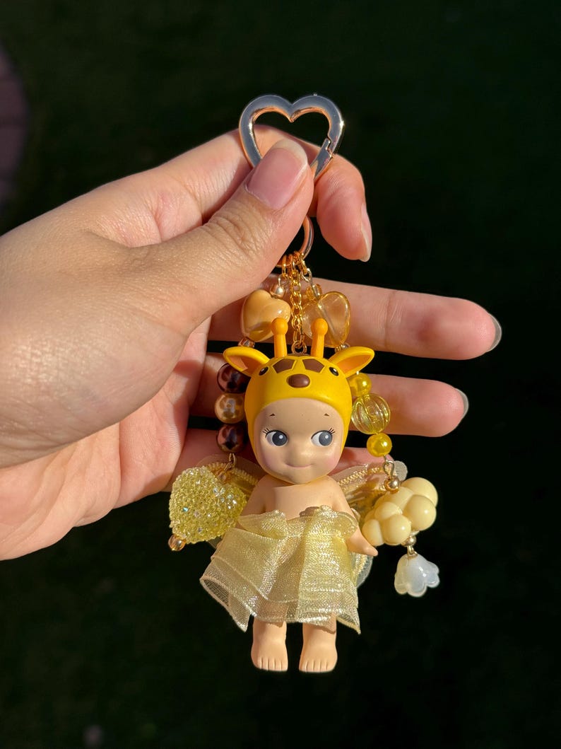 Puede incluir: Un llavero amarillo y dorado con una peque&ntilde;a mu&ntilde;eca vestida de jirafa. La mu&ntilde;eca tiene una capucha de jirafa amarilla, una falda amarilla transparente y peque&ntilde;as alas amarillas. El llavero incluye cuentas doradas y un cierre en forma de coraz&oacute;n.