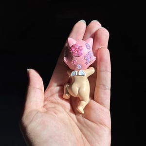 May include: A small figurine of a cat-like creature with a light pink head adorned with iridescent floral-shaped crystals. The figure has a tan-colored body, small white wings on its back, and is depicted in a crawling position with its head turned.